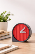 Red clock with black frame on a wooden surface, books, and a plant in the background