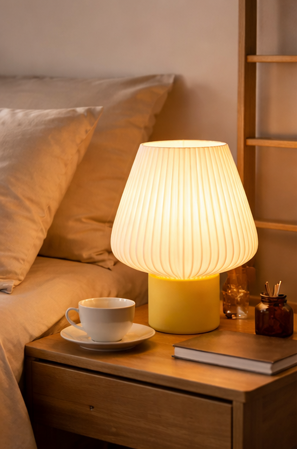 Yellow table lamp on a bedside table with a cup and saucer, book, and glasses.